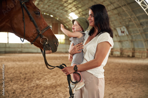 Happy kid is touching the animal. Gorgeous woman with her little baby son is with horse indoors