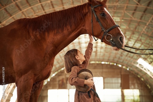 Side view. Cute little girl is with horse indoors