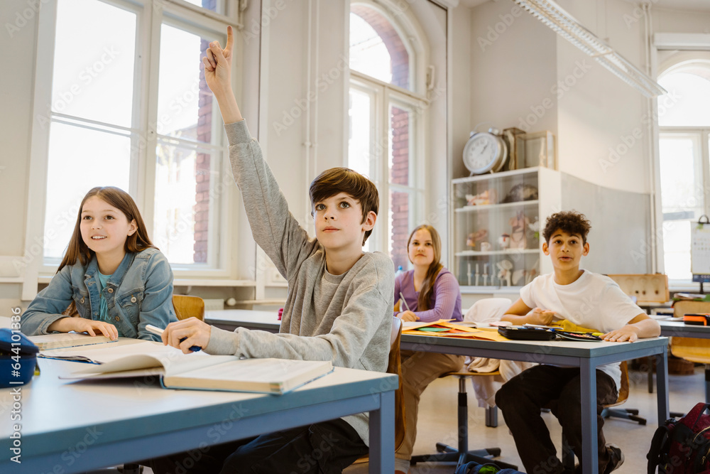 Boy raising hand while sitting with female friend at desk in classroom Boy raising hand while sitting with female friend at desk in classroom