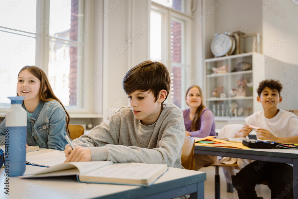 Schoolboy writing in book while sitting at desk with friends in classroom Schoolboy writing in book while sitting at desk with friends in classroom