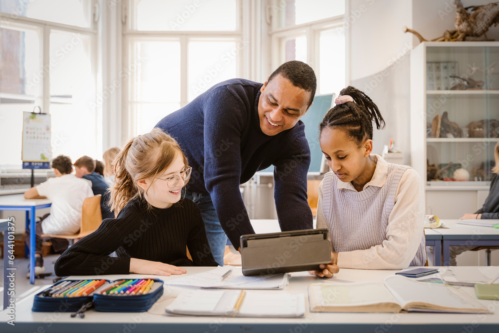Smiling male teacher doing E-learning with female students in classroom Smiling male teacher doing E-learning with female students in classroom