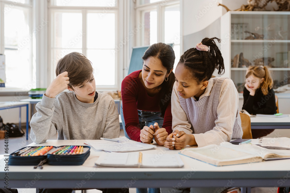 Teacher discussing with male and female students sitting at desk in classroom Teacher discussing with male and female students sitting at desk in classroom