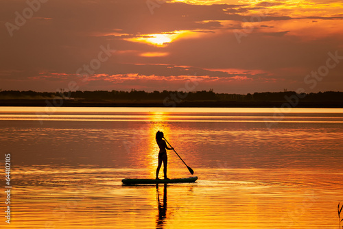 woman stands on a sup board with a paddle on the rowing channel against the background of a yellow sunset. Active water sports. A sexy woman is engaged in rowing.