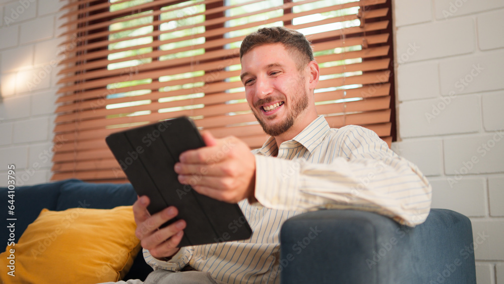 Caucasian man, Bearded smiling using touch screen digital tablet. While sitting on sofa in living room at home.