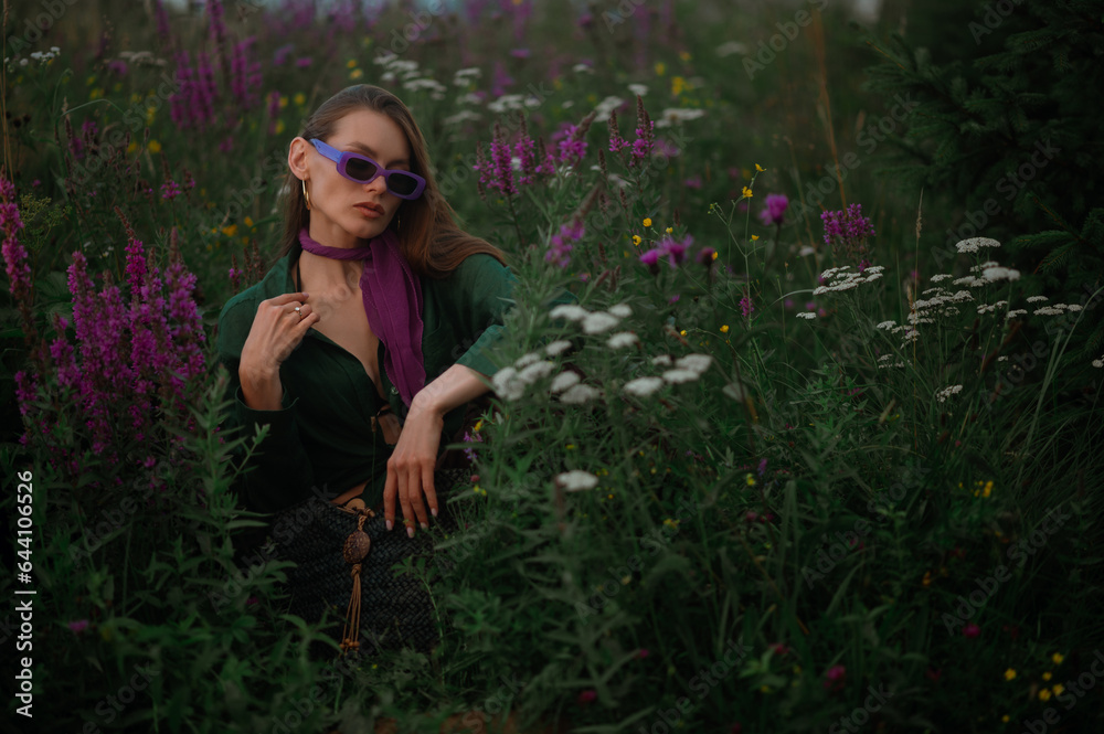 Fashionable confident woman wearing trendy purple sunglasses, neck scarf, hoop earrings, green linen shirt, sitting, posing in summer field. Copy, empty space for text Fashionable confident woman wearing trendy purple sunglasses, neck scarf, hoop earrings, green linen shirt, sitting, posing in summer field. Copy, empty space for text