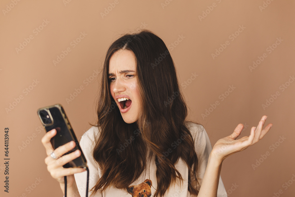 Mad and shock young brunette beautiful woman using mobile phone isolated on beige background. Yelling unhappy woman hold mobile phone. Disappointed sad upset lady horrified impressed news. WTF. Oh no. Mad and shock young brunette beautiful woman using mobile phone isolated on beige background. Yelling unhappy woman hold mobile phone. Disappointed sad upset lady horrified impressed news. WTF. Oh no.