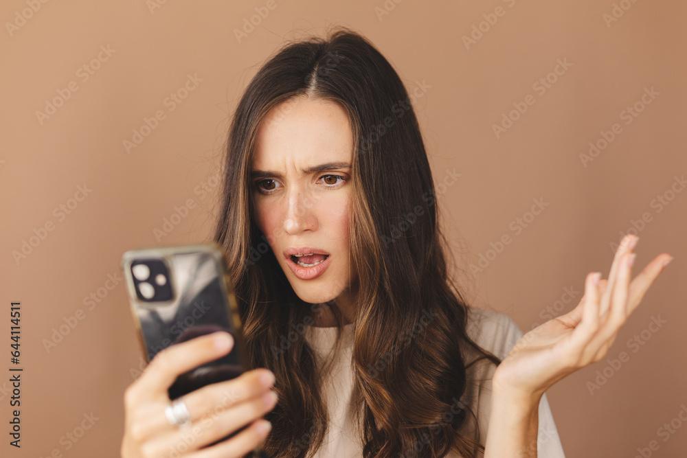 Mad and shock young brunette beautiful woman using mobile phone isolated on beige background. Yelling unhappy woman hold mobile phone. Disappointed sad upset lady horrified impressed news. WTF. Oh no. Mad and shock young brunette beautiful woman using mobile phone isolated on beige background. Yelling unhappy woman hold mobile phone. Disappointed sad upset lady horrified impressed news. WTF. Oh no.