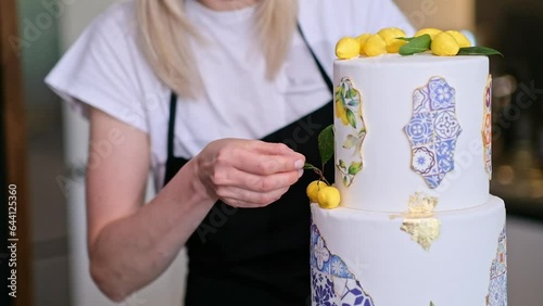 Satisfied housewife putting decorative green leaf near yellow lemons on top of two-tiered cake standing in kitchen small business of organic sugar free confectionery production at home photo