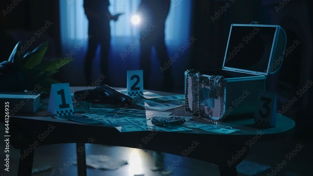 Closeup shot of the table with evidence numbers in the dark apartment room, police officers standing in the background.