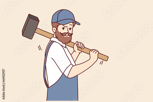 Bearded man with hammer works in auto repair shop or at construction site, dressed in overalls with cap. Brutal worker holding hammer offering to demolish old house and build modern mansion