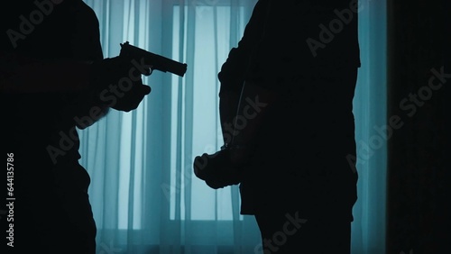 The arrest of a robber in a dark apartment lit with blue light. Side view of a criminal in black clothes with handcuffed hands behind his back. A male police officer holds the villain at gunpoint.