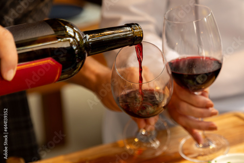 Close em garrafa de vinho tinto sendo servido em duas taças seguradas por casal em fundo desfocado.
