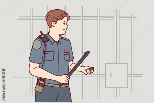 Strict man prison guard with rubber club in hand walks near cell for criminals and keeps order in jail. Guy works as prison guard, dressed in uniform of police officer or employee security service