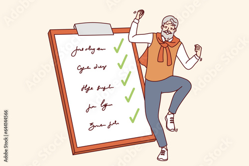 Elderly man completed all tasks and dancing near giant clipboard with list of goals and checkmarks. Gray-haired happy pensioner celebrating achievement of set goals after retirement