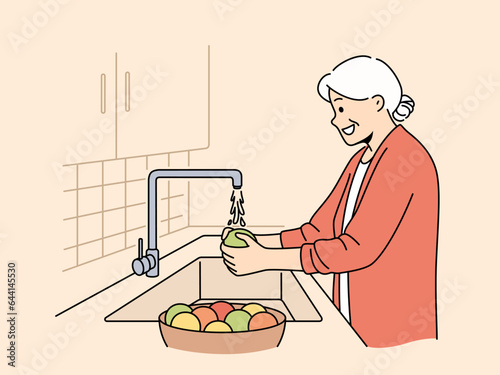 Elderly woman washes fruits in sink, standing in kitchen and observing rules of hygiene to avoid infection. Grandmother washes vegetables before preparing dinner, following recommendations of doctors