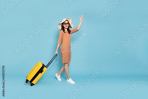 Happy young Asian woman traveler walking and drag luggage isolated on blue sky background, Tourist girl having cheerful holiday trip concept, Full body composition
