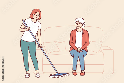 Caretaker from nursing home is mopping floor and taking care of elderly woman sitting on couch. Gray-haired female pensioner with smile watches caretaker cleaning room to maintain cleanliness