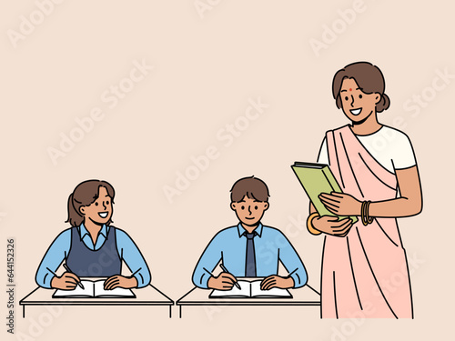 Indian woman teacher works in primary school, standing near children sitting at desks in classroom. Girl teacher from india, makes career in education and gives kids new knowledge.