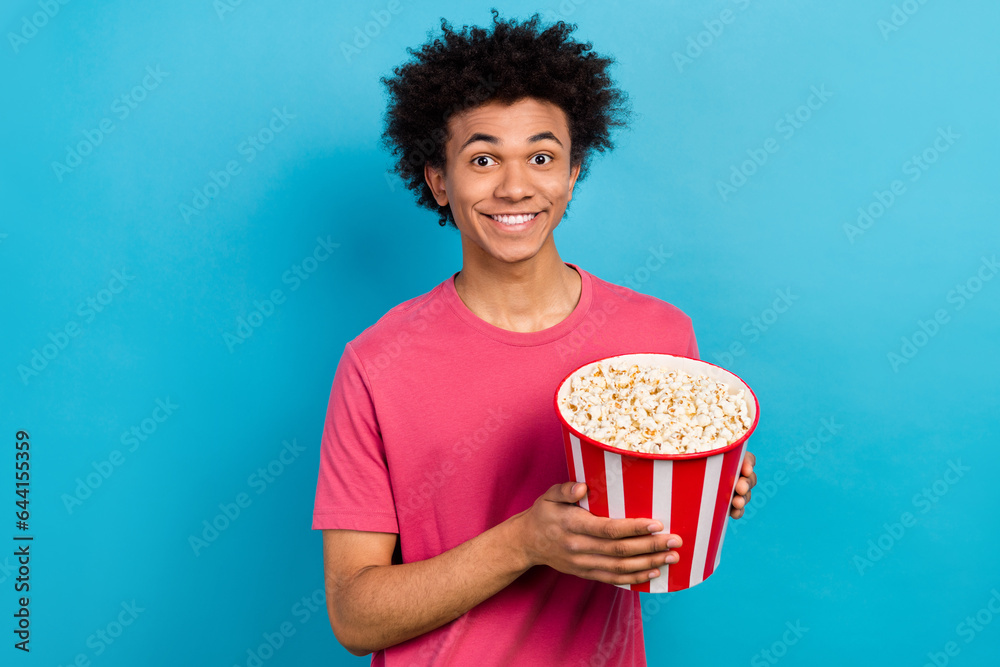 Portrait of cheerful guy with perming coiffure dressed stylish t-shirt hands hold pop corn basket isolated on blue color background Portrait of cheerful guy with perming coiffure dressed stylish t-shirt hands hold pop corn basket isolated on blue color background