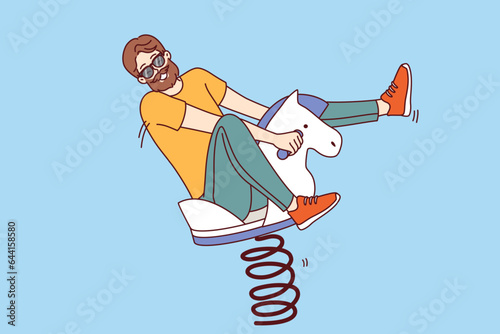 Playful man sits on wooden rocking horse and enjoys children games, behaves like child of adolescence. Bearded man in sunglasses looks at screen, is childish and does not want to grow up