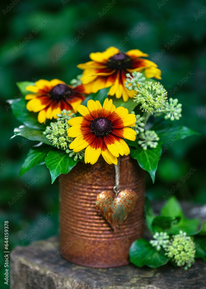Obraz premium Flower arrangement with yellow and orange globular or Black Eyed Susan flowers and ivy blossom in rusty tins with rustic heart in the summer garden. Gardening concept. Selective focus.