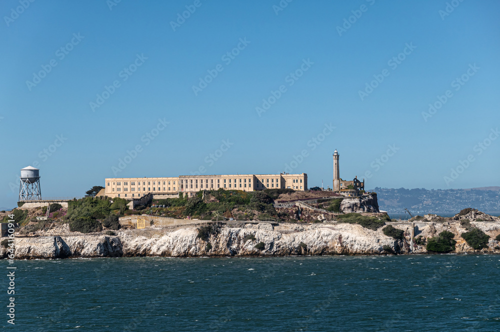 San Francisco, CA, USA - July 13, 2023: West shore of Alcatraz Island showing the main building, water tower and lighthouse. Shoreline covered by white guano under blue sky