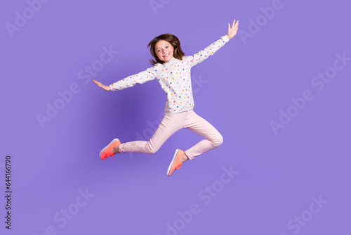 Full length photo of carefree pretty little girl dressed dotted pullover arms sides jumping high isolated purple color background