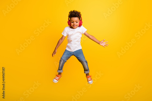 Full length photo of positive cheerful little boy dressed white t-shirt jumping high enjoying songs earphones isolated yellow color background