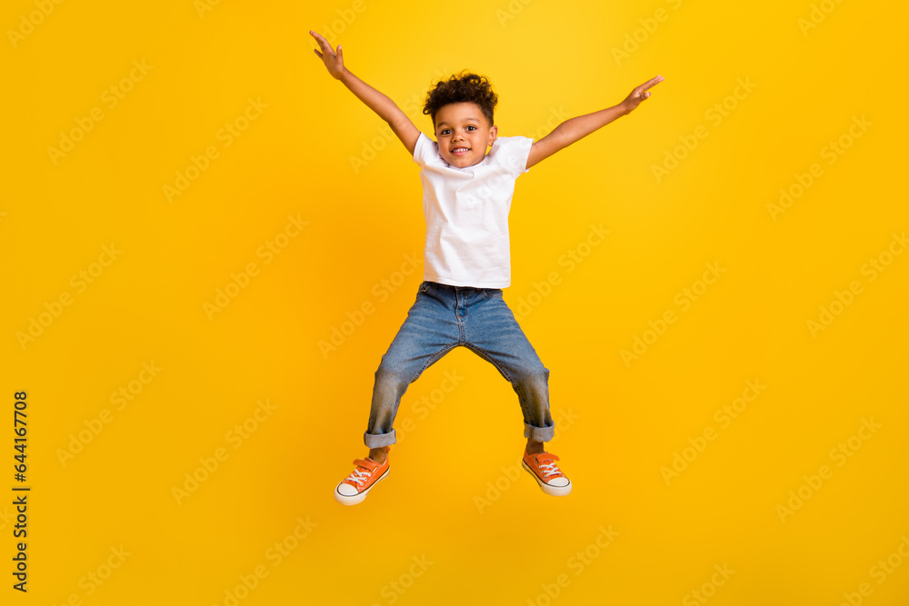 Full length photo of sweet excited little boy dressed white t-shirt jumping high arms sides isolated yellow color background Full length photo of sweet excited little boy dressed white t-shirt jumping high arms sides isolated yellow color background