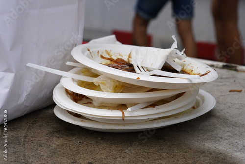 White plastic plates are stacked on one another. There are rests of the food on the  plates as well as some pieces of white plastic cutlery. On the defocused background there are passers by. 