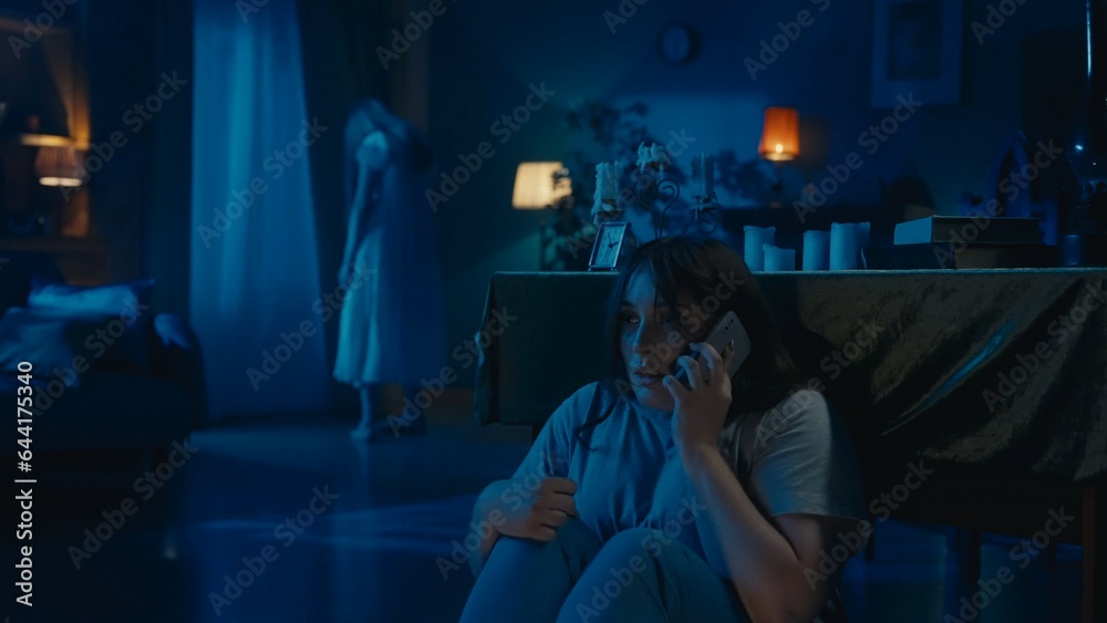 Shot of a young woman sitting, hiding behind the table from from an otherwordly creature, entity, ghost. Woman is calling somebody. Shot of a young woman sitting, hiding behind the table from from an otherwordly creature, entity, ghost. Woman is calling somebody.