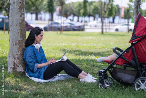 Young woman with headphones working on tablet computer while baby sleeping in buggy on summer park lawn mother surfing internet via device walking with infant child photo