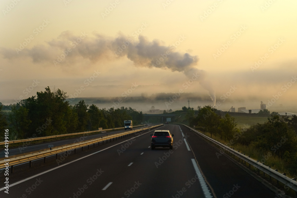Near the motorway there is an industrial enterprise from the chimney of which there is thick smoke, in the rays of the rising sun