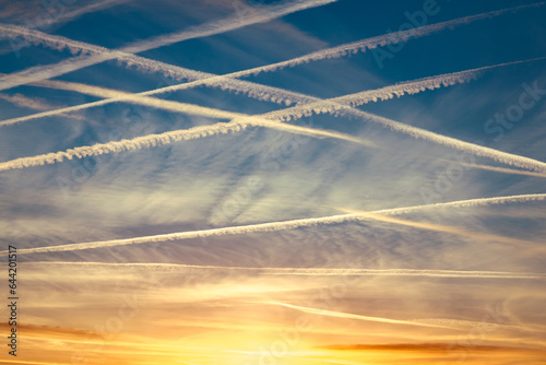Around 10 airplane vapor trails in the sky in a diagonal pattern against a dark blue sky at sunset with yellow and orange clouds on the horizon.
