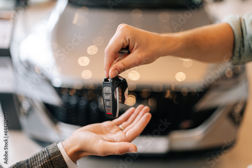 Close-up hands of unrecognizable dealer male giving new car key to customer man in business suit on blurred background of new auto in dealership office. Concept of choosing and buying car at showroom.