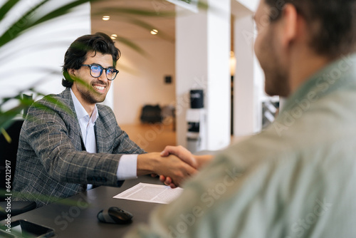Selective focus of smiling real estate broker in business suit present and advise young man client on decision to sign insurance contract. Happy customer signing purchase agreement and shaking hands. photo