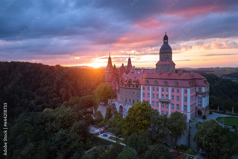 An evening aerial view of the red sunlit Ksiaz Castle, Schloss Fürstenstein, a beautiful castle standing on a rock surrounded by forest against a dramatic sky. Poland.