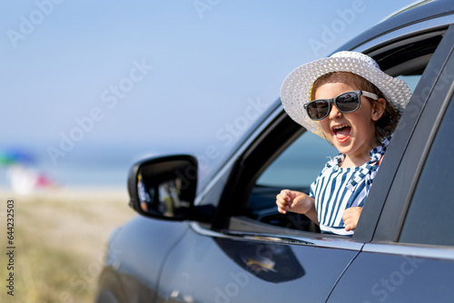 Cute toddler girl wearing sunhat and sunglasses looking through open window from driver seat at the seaside. On the road summer trip to the beach. Space for copy.