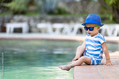 Fototapeta Naklejka Na Ścianę i Meble -  Baby boy wearing sunhat and sunglasses sitting at the edge of swimming pool. Summer relaxation and childhood concept.