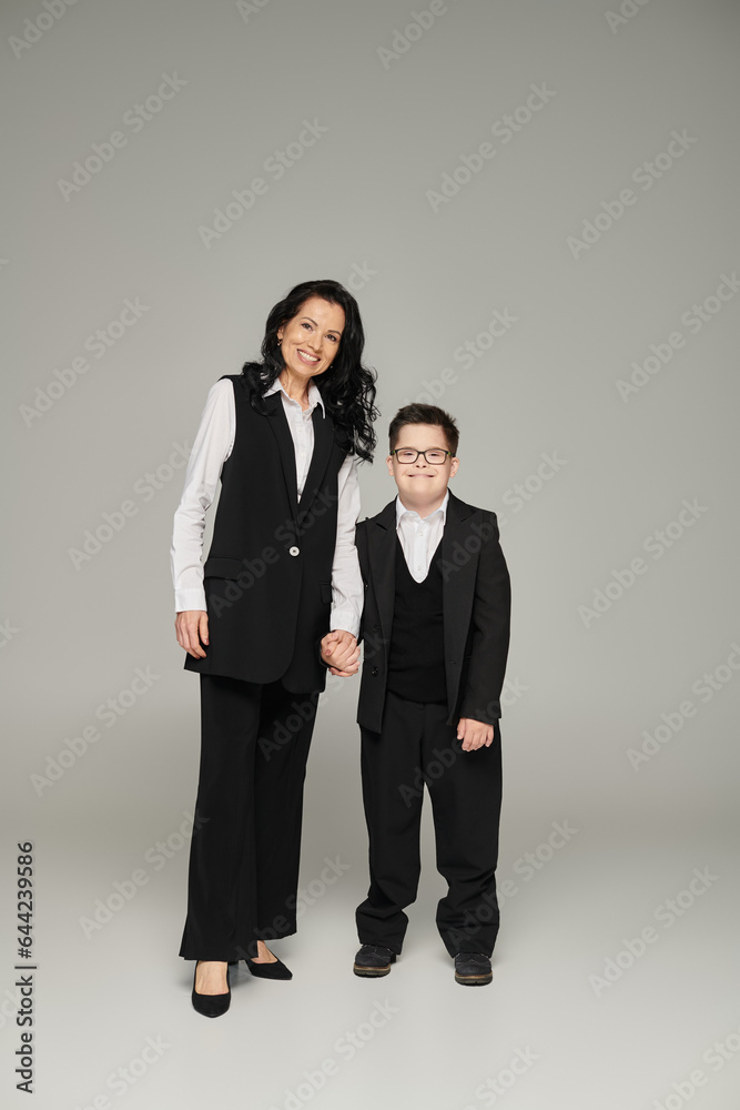 happy woman in formal wear holding hand of son with down syndrome in school uniform on grey happy woman in formal wear holding hand of son with down syndrome in school uniform on grey