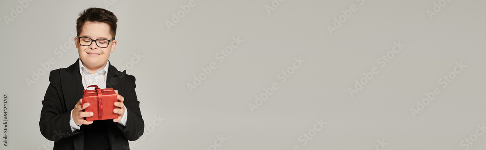 joyful kid with down syndrome in school uniform and eyeglasses holding red gift box on grey, banner joyful kid with down syndrome in school uniform and eyeglasses holding red gift box on grey, banner