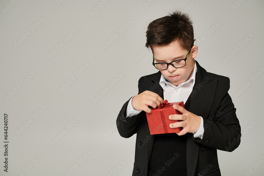 boy with down syndrome in school uniform and eyeglasses opening gift box with red ribbon on grey boy with down syndrome in school uniform and eyeglasses opening gift box with red ribbon on grey
