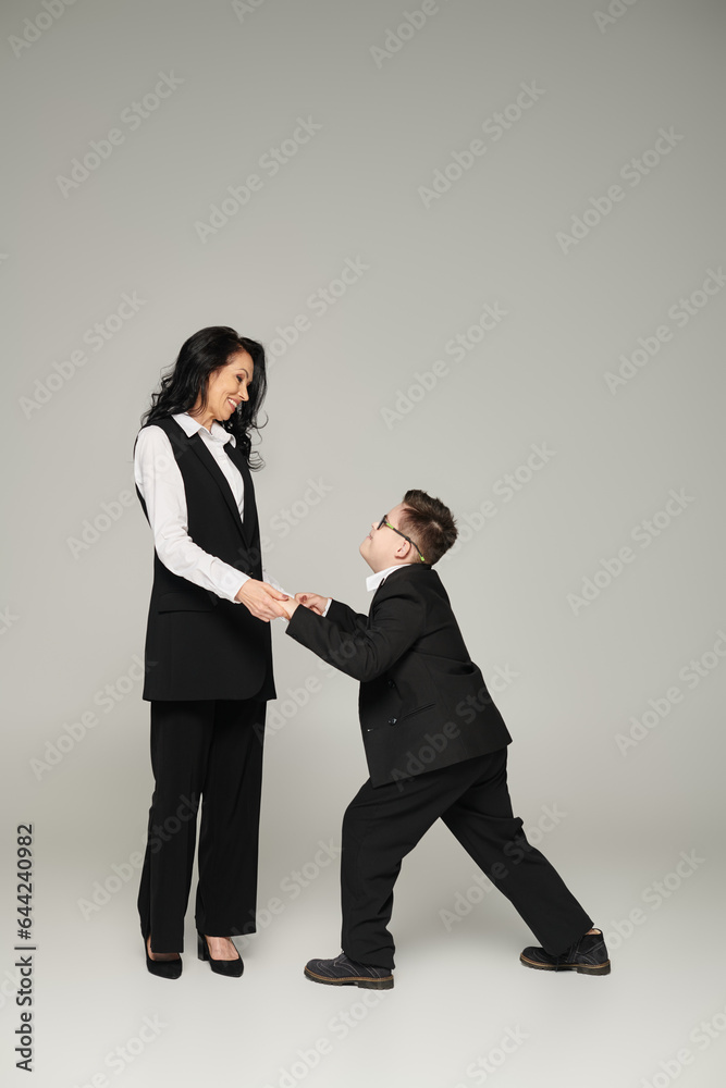 schoolboy with mental disability holding hands of happy mother in formal wear on grey, full length schoolboy with mental disability holding hands of happy mother in formal wear on grey, full length
