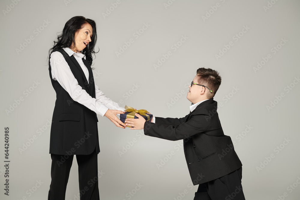 kid with down syndrome giving present to amazed mother in elegant outfit on grey, unique family kid with down syndrome giving present to amazed mother in elegant outfit on grey, unique family