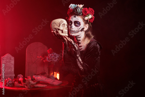 Santa muerte model holding skull in studio, acting scary and horror to celebrate mexican halloween. Beautiful woman in festival costume with body art, looking like goddess of death.