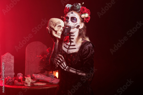 La cavalera catrina acting creepy with skull in hand, holding black roses and wearing scary make up. Looking like santa muerte with flowers crown and body art on holy mexican celebration.