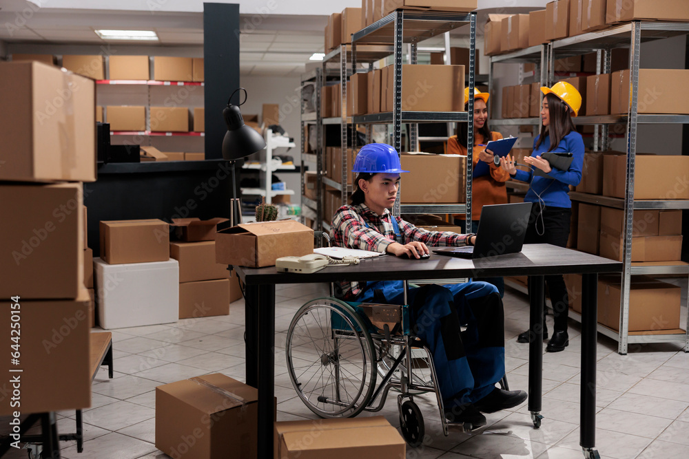 Young asian storehouse employee with physical disability managing delivery operations on laptop. Freight distribution center worker in wheelchair doing desk work in warehouse