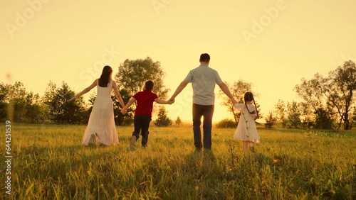 Happy family run forward on green grass park. Mom, dad, daughter, son holding hands running across field. Parents children hold hands run towards sunset. Mother father kid play in nature. Young family