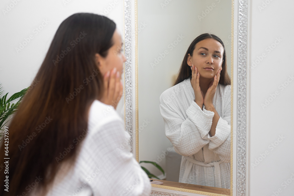 Attractive brunette woman in gown looking in mirror in bathroom lady checking facial skin condition standing by dressing table at home woman with healthy skin near mirror Attractive brunette woman in gown looking in mirror in bathroom lady checking facial skin condition standing by dressing table at home woman with healthy skin near mirror
