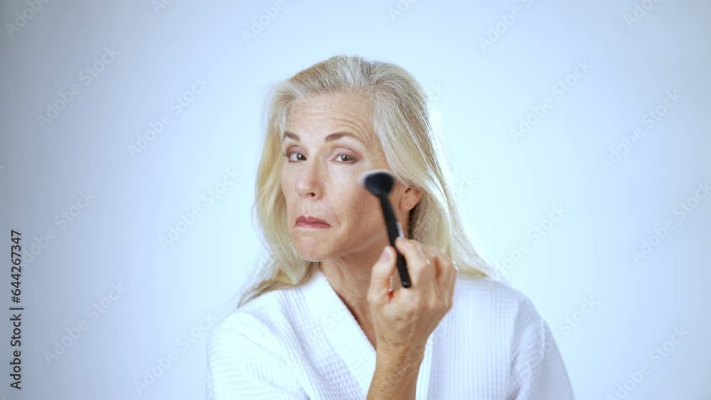 Portrait of mature blond and gray hair woman looking in the mirror, puts powder on her face with brush Portrait of mature blond and gray hair woman looking in the mirror, puts powder on her face with brush